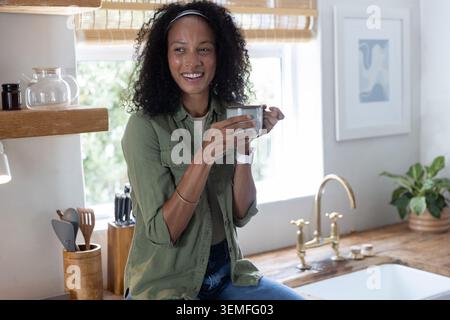 Woman sitting on kitchen countertop beside brass faucet over white sink holding ceramic mug smiling Stock Photo