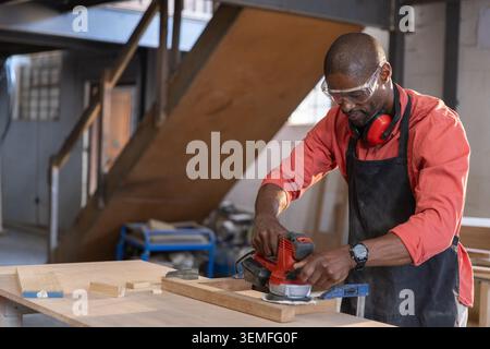 Senior African American man using electric sander sanding wooden plank in workshop, copy space Stock Photo