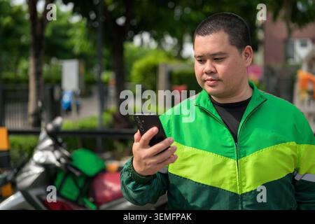A high-resolution horizontal portrait of a plus-size Thai male delivery rider or courier actively using a black smartphone device outdoors. The subjec Stock Photo