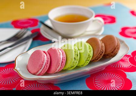 Assorted colorful macarons arranged on a plate beside a cup of tea and fork on a bright patterned table in Japan. Stock Photo