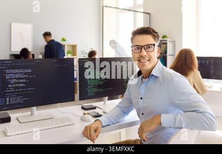 Confident programmer posing by code monitors at desk in bright modern office Stock Photo
