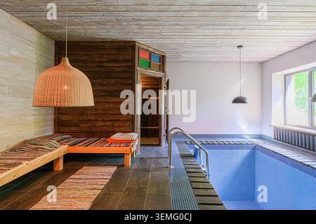 Indoor pool room with a sauna, lounge area with a woven lamp, and a tiled pool with a ladder. Rustic and modern elements blend. Stock Photo