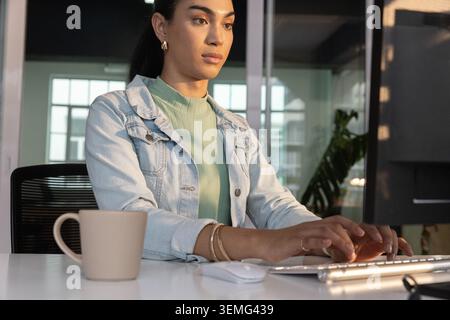 Woman sitting at white desk in office typing on slim silver keyboard with beige mug Stock Photo