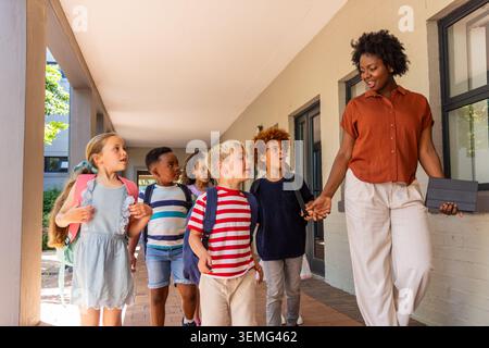 Diverse children wearing backpacks walking along school corridor with female teacher holding tablet. Group, pupils, education, outdoor, sunlight, gree Stock Photo
