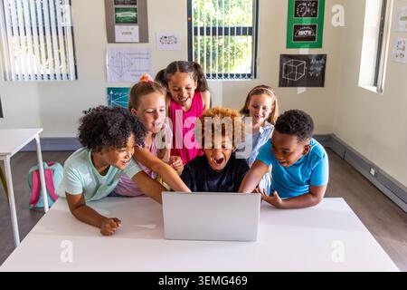Diverse children leaning around open laptop in classroom, pointing at laptop screen and backpack Stock Photo