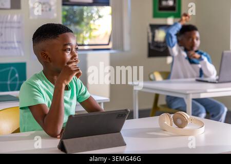 African American school-age boys sitting at desks in classroom, sharing focus on tablet and laptop Stock Photo