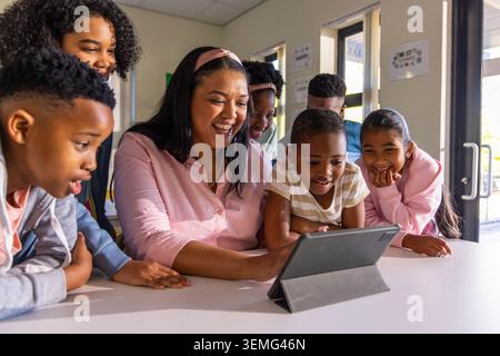 Female teacher in pink blouse and headband guiding diverse children around tablet at school table Stock Photo