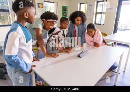 Diverse children and teacher leaning in around child wearing VR headset in classroom using tablet. Education, learning, collaboration, technology, sch Stock Photo