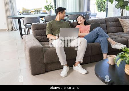 Diverse couple sitting on gray sectional sofa smiling while using laptops in living room Stock Photo