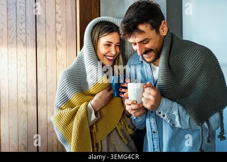 Happy young couple with hot drinks cuddling under warm blanket at home Stock Photo