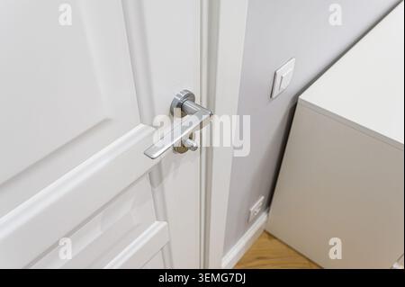 A close-up of a white door with a brushed metal handle. A light switch is visible on the wall beside the door, and part of a white cabinet is in the b Stock Photo