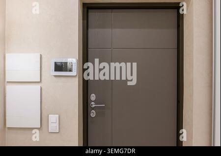 Interior shot of a brown door in a textured beige wall.  Intercom, white panels and a light switch are mounted on the wall next to the door. Stock Photo