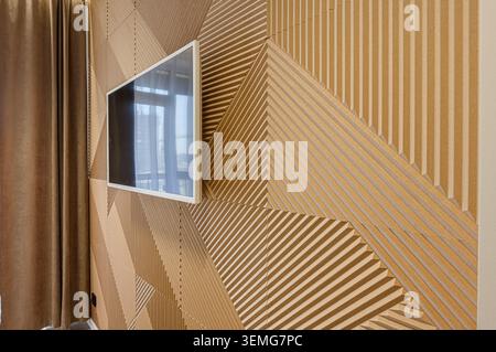 Modern interior with a decorative wooden wall panel featuring geometric patterns. A flat-screen TV hangs on the wall, reflecting a window. Brown curta Stock Photo