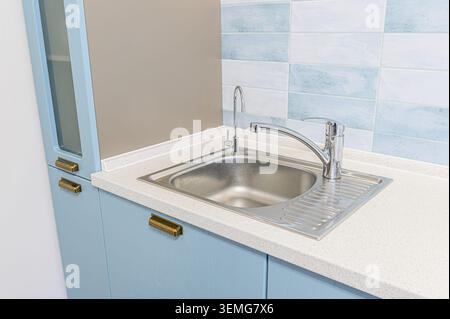 kitchen sink with a faucet and tap on a light countertop. Cabinets are blue with bronze handles, backsplash with white and blue tiles Stock Photo