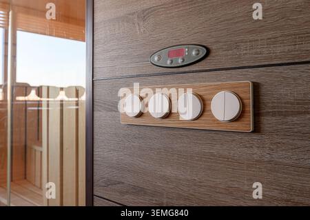 control panel on a wooden sauna wall features a digital display and four round switches. The sauna interior is visible through a glass door. Stock Photo