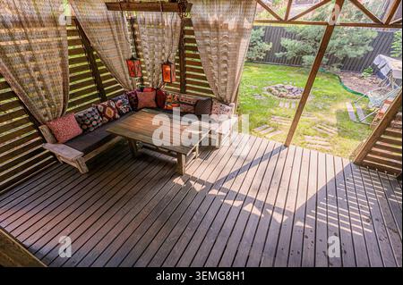 cozy wooden gazebo features draped curtains, colorful pillows on a built-in bench, and a wooden table. Outside, a green lawn extends. Stock Photo