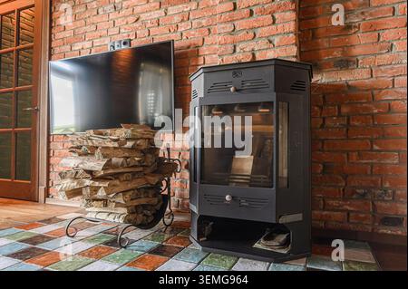 Cozy room with brick wall, TV above stacked firewood, and a black stove. Tiled floor adds a colorful touch. Stock Photo