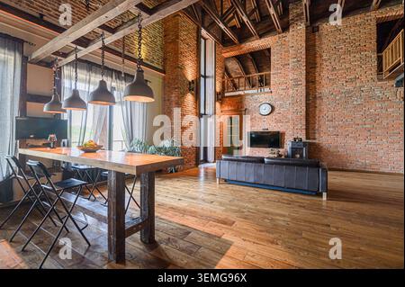 spacious loft with exposed brick walls, wooden floors, and a high ceiling. A long table with industrial-style lamps dominates the foreground Stock Photo