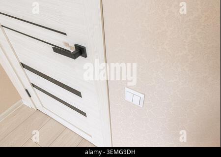 close-up features a modern white door with black accents, a black handle, light switch, and a textured beige wall. The floor is light wood-toned. Stock Photo