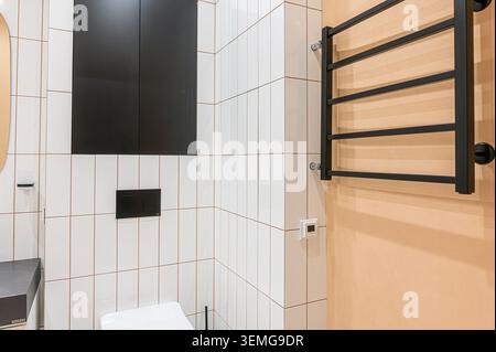 modern bathroom featuring white tiled walls with dark grout, a black cabinet, toilet flush plate, and a black towel warmer against a peach wall Stock Photo
