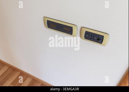 Two beige electrical panels are mounted on a white wall above a wooden floor. One panel has a digital display, and the other has outlets and a dimmer Stock Photo