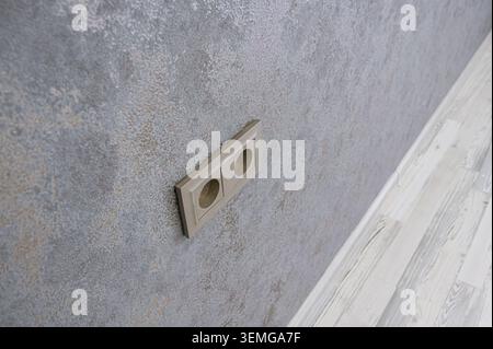 close-up shows a double electrical outlet on a textured gray wall, with a view of the light-colored wooden floor near the wall's edge. Stock Photo
