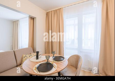 Cozy dining area with round table, set for two, next to a beige sofa and bright window with sheer curtains Stock Photo