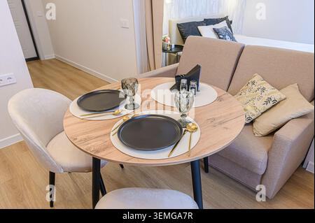 Cozy dining area with a round table set for two, featuring elegant gray plates and glassware, next to a comfortable couch and stylish bedroom Stock Photo