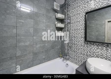 Modern bathroom with grey and mosaic tiles, a white bathtub, a shower, and a white sink on a dark gray vanity Stock Photo