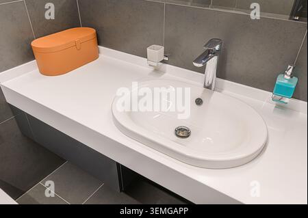 Modern bathroom sink with white countertop, chrome faucet, and orange storage box. Gray tile background. Stock Photo