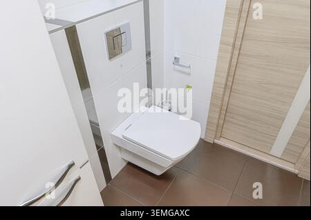 modern bathroom features a wall-mounted toilet, white tiles with accents, a light wood door, and brown textured floor tiles Stock Photo