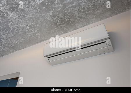 white air conditioner unit is mounted on a white wall below a textured grey ceiling. Minimalist and modern aesthetic Stock Photo