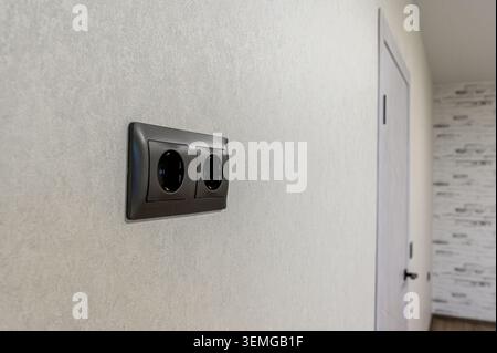 close-up shows a dark double electrical outlet mounted on a textured, light-colored wall. A partially visible door is in the background. Stock Photo