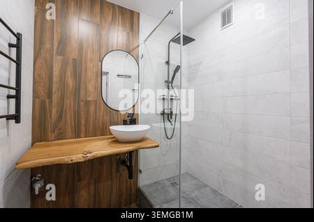 Modern bathroom with wood accents. Features a glass-enclosed shower, oval mirror, and unique wood countertop with a white sink Stock Photo