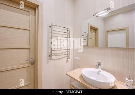 Bathroom view featuring a light wood door, chrome towel warmer, oval sink on wood counter, mirror, and neutral striped wall tiles. Stock Photo