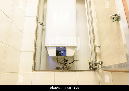 white water heater is mounted in a tiled enclosure, with an open cabinet door revealing its display panel and plumbing connections Stock Photo