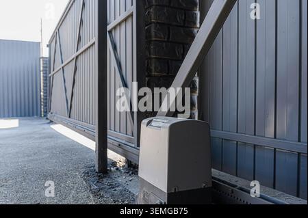 low-angle shot of an automated gray metal gate with a brick pillar, focusing on the motor mechanism. Stock Photo