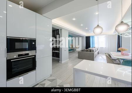 Modern interior with white cabinets and appliances, open living area, pendant lights, and a gray sofa. Clean lines and a bright, airy atmosphere. Stock Photo