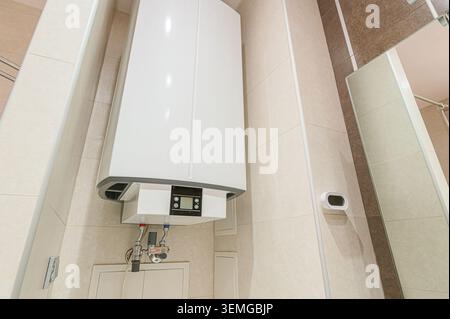 water heater is mounted on a tiled wall in a bathroom. The water heater is white, and the bathroom features beige tiles and modern fixtures Stock Photo