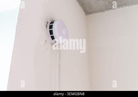 smoke detector is mounted on a white wall in a corner. The wall has some stains and the ceiling is visible Stock Photo