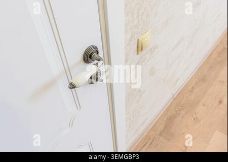 Close-up of a white paneled door with an antique-style brass handle and a white porcelain grip. The wall next to it features a light-colored switch an Stock Photo