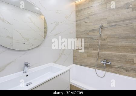 modern bathroom featuring a white sink, round mirror, bathtub, and shower. Walls are tiled with marble and wood-patterned tiles. Stock Photo