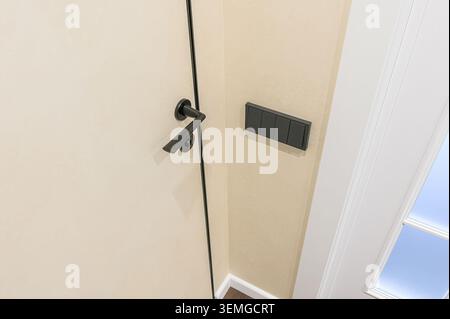 close-up shot of a door with a black handle and frame, next to a wall with a gray light switch panel, adjacent to a white framed window or doorway. Stock Photo
