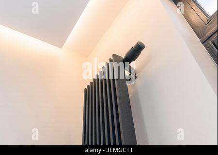 modern black radiator mounted on a light-colored wall is viewed from below. A cylindrical valve is affixed to the top. The room features ambient light Stock Photo