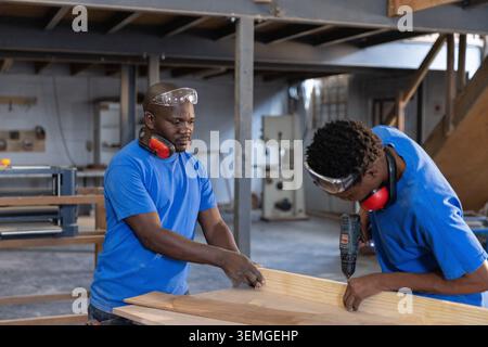 African American male coworkers securing plank using drill in workshop wearing goggles and muffs Stock Photo