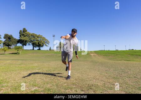 Mid-adult man making throwing motion on park field wearing black cap, compression sleeve Stock Photo