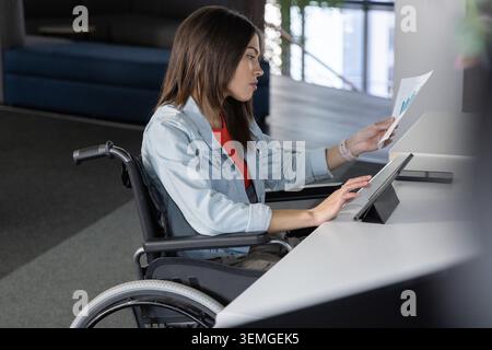 Woman in wheelchair reviewing charts and using tablet at coworking white desk with smartphone Stock Photo