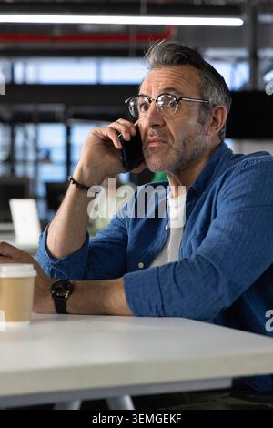 Middle-aged man sitting at desk in office, holding phone near coffee cup wearing round glasses Stock Photo