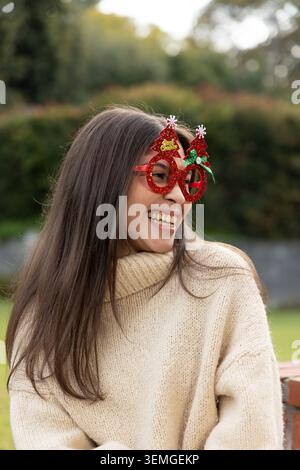 Woman in early 20s laughing in garden wearing cream-turtleneck and red-glitter-tree-glasses on wall Stock Photo