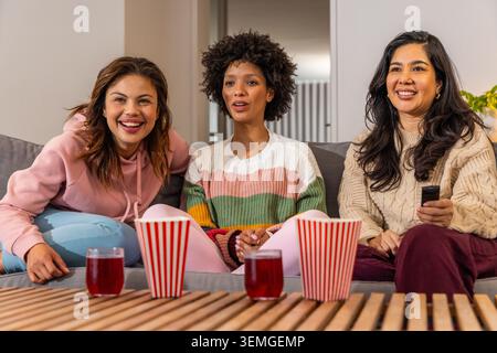Diverse female friends sitting on gray sofa in living room watching TV with popcorn, drinks, remote Stock Photo